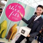 The picture shows a smiling presenter holding a box of shoes, he is standing on front of a promotional poster advertising the fun fashion game, If The Shoe Fits