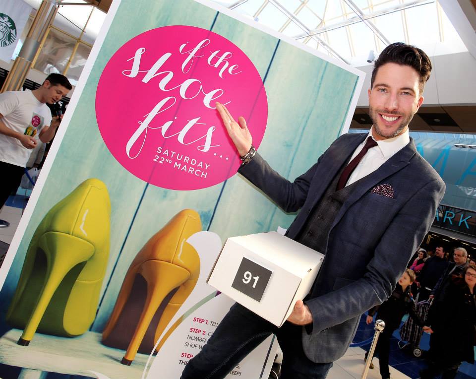 The picture shows a smiling presenter holding a box of shoes, he is standing on front of a promotional poster advertising the fun fashion game, If The Shoe Fits