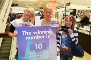 The picture depicts 3 smiling ladies in a retail store. One lady, the she store manager, holds the prize number, the other lady holds the winning ticket with the matching store number, and her daughter holds their prize, a gift card from the retailer.