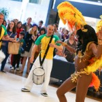 The picture shows a dancer in colourful carnival costume dancing to a Samba beat provided by a band of drummers. The smiling crowd in the background are enjoying the show.