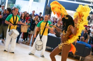 The picture shows a dancer in colourful carnival costume dancing to a Samba beat provided by a band of drummers. The smiling crowd in the background are enjoying the show.