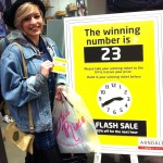 The pictures shows a smiling female student holding the prize winning ticket number, she is standing next to the in-store display of the matching number displayed in store