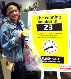 The pictures shows a smiling female student holding the prize winning ticket number, she is standing next to the in-store display of the matching number displayed in store