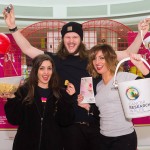 Group of three charity workers celebrate big donations to Cancer Research Wales by holding up collection buckets and cheering. They are stood in front of the giant Valentine heart installation at the Love Lock-In retail event Cardiff