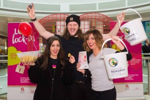 Group of three charity workers celebrate big donations to Cancer Research Wales by holding up collection buckets and cheering. They are stood in front of the giant Valentine heart installation at the Love Lock-In retail event Cardiff