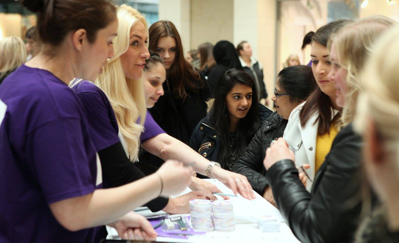 A crowd of women talk to staff at a registration desk, they are signing up to participate in a beauty event at a busy shopping centre.