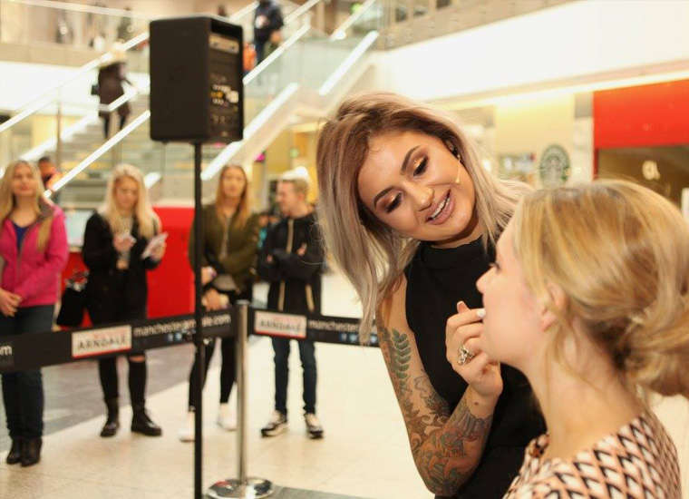 Makeup artist applying makeup to young woman's face during live beauty demonstration at shopping mall. In the background there are spectators.