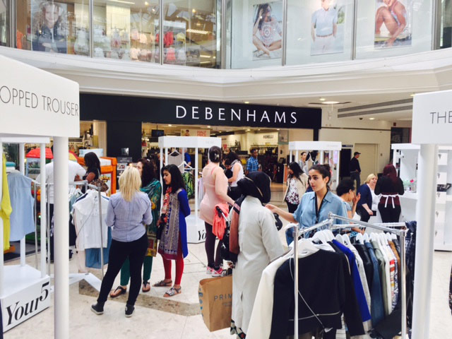 View of a busy shopping centre with temporary display stands showing items of clothing are installed on the mall. There are crowds of shoppers milling about, talking to brand associates.