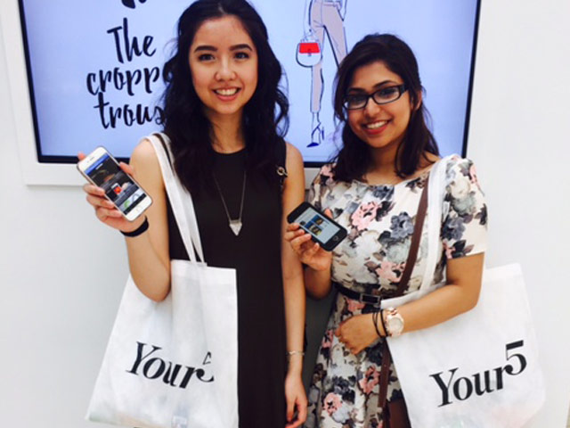Two young women hold up their smart phones towards camera to show they have downloaded an app for the Your 5 retail shopping event. They stand in front of the event campaign branding and each has a branded shopping bag.