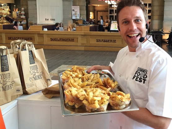 A smiling chef presents a tray of restaurant food samples towards the camera. The restaurant can be seen in the background.