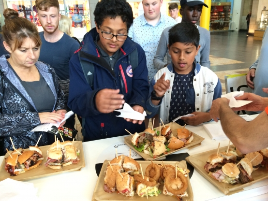 A group of people gather around a table filled with a variety of food samples, they are eager to try the dishes on offer.