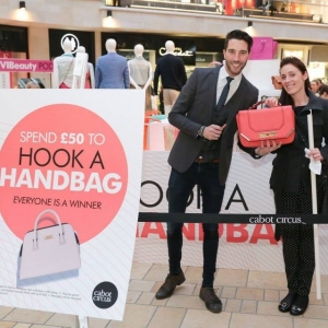 The experience economy - A woman poses for the camera alongside the presenter of a live experiential fashion event taking place at a shopping mall. She has won a prize, a designer handbag, and holds it up towards the camera to show it off.