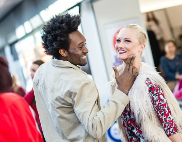 Male model tenderly touches the face of female model, both are smiling and dressed in stylish clothing.