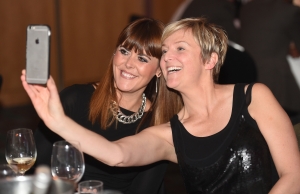 Two well dressed women are sat at a table during an evening awards ceremony, they are smiling into their smartphone taking selfies.