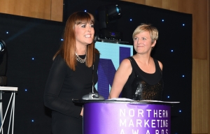 Smartly dresssed woman stood at podium, talking into microphone, making acceptance speech at Northern Marketing Awards. A second woman is standing off to the right, looking at the speaker and smiling.