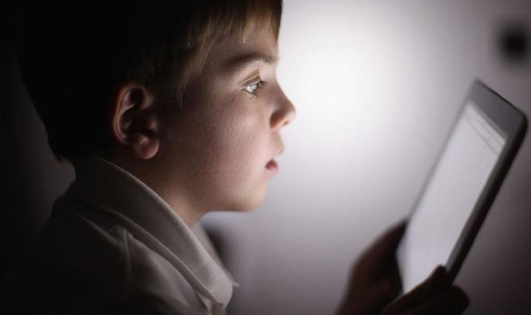 Experiential learning. Little boy sat in dark room looking at tablet. The light from the screen illuminates his face.