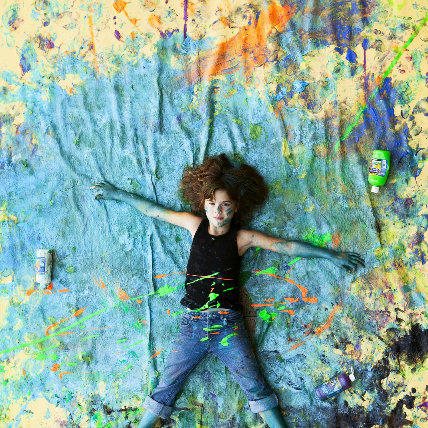 Experiential learning. Photo , shot from above, shows little girl lying on a paint splattered canvas. She is covered in paint and is loking up towards camera, smiling.